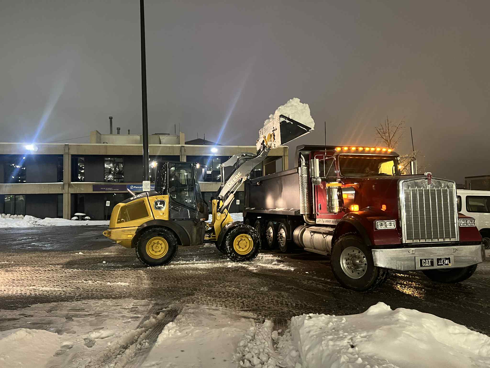 Front loader dumps snow into a red dump truck during nighttime commercial snow removal in an Ames, Iowa business parking lot.