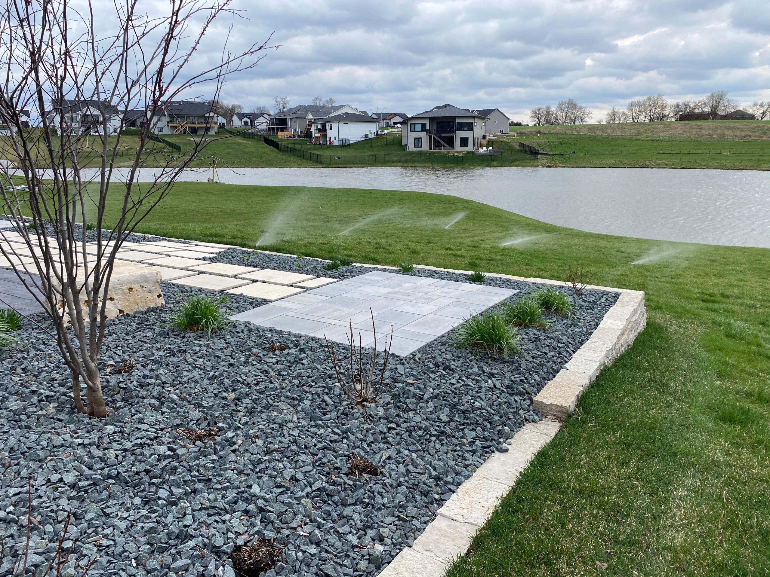 Lawn irrigation system watering a green yard near a lake, with a landscaped stone pathway and modern houses in the background.